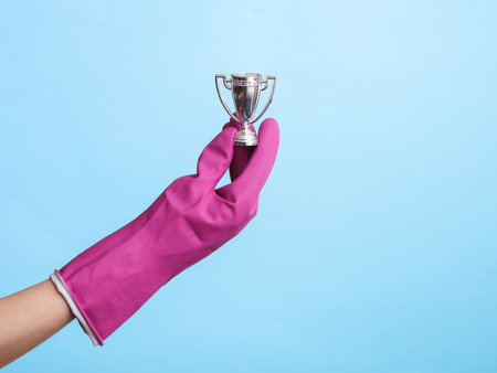 Hand in purple rubber cleaning glove holding cup trophy on blue background. House cleaning and housekeeping conceptの写真素材