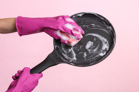 Hands in rubber gloves wash frying pan with foam on pink background