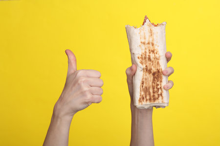 Woman's hand holds an appetizing bitten shawarma and shows a thumbs up on a yellow backgroundの写真素材