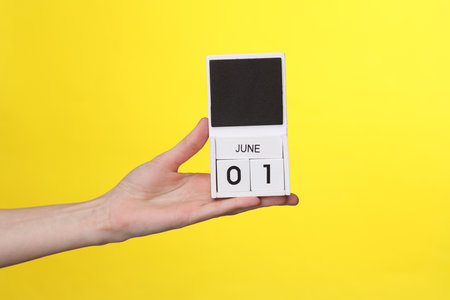 Woman's hand holds wooden calendar with date june 01 on a yellow backgroundの写真素材