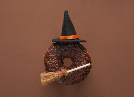 Chocolate donut with witch's broom and hat on brown background. Halloween concept. Trick or Treat