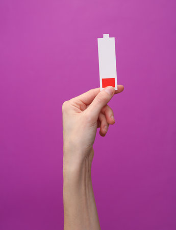 Woman's hand holding a paper-cut icon of a battery with low charge level on purple background