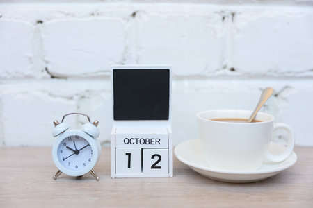 Wooden calendar with date October 12 and alarm clock with coffee cup on table against brick wall...