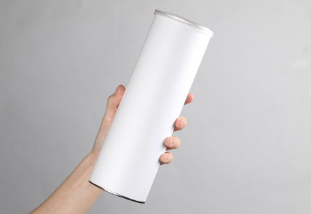 Woman's hand holds a cylindrical tin can with a white empty label on gray background. Template for designの写真素材