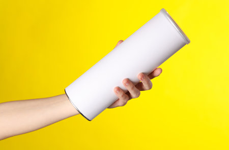 Woman's hand holds a cylindrical tin can with a white empty label on yellow background. Template for designの写真素材