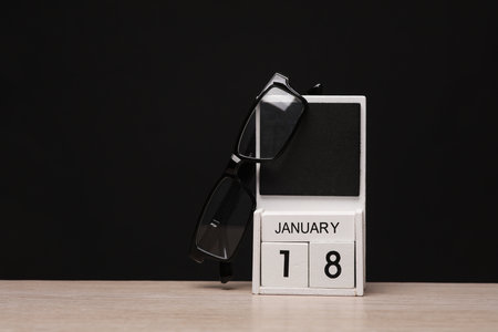 White wooden block monthly calendar with the date january 18 and eyeglasses on the table, black blackbackground. Planningの写真素材