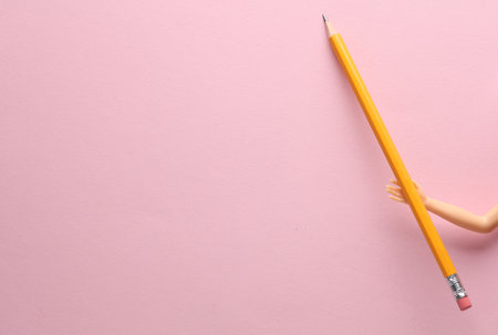 The doll's hand holds a pencil on a pink background. minimal layoutの写真素材