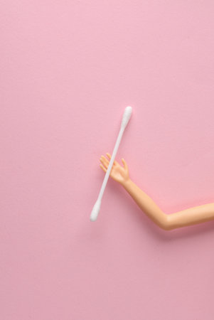 The doll's hand holds a cotton swab on a pink background. Ear hygiene concept