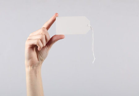 Female hand holds a white blank price tag on gray background. sale conceptの写真素材