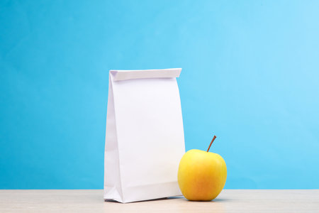 White lunch pack and apple on the table, blue background. Snack to school. Template for designの写真素材