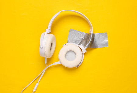 White stereo headphones fixed with adhesive tape on yellow background. Conceptual pop, contemporary artの写真素材