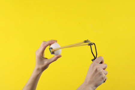 Hands holding slingshot with egg on a yellow backgroundの写真素材