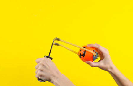 Hands holding slingshot with toy Jack pumpkin bucket on a yellow background. Halloween theme, trick or treatの写真素材