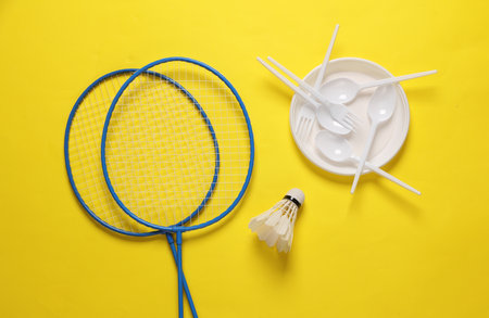 picnic. Badminton racket with shuttlecock and plastic plate with spoons and forks on yellow background with shadowの写真素材