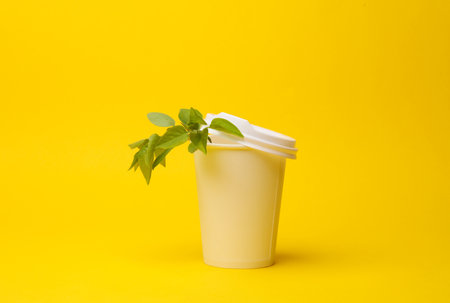 Disposable cardboard coffee cup with a green leaf branch on yellow background. Eco conceptの写真素材
