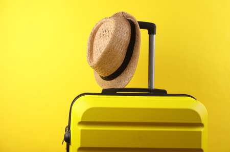 Yellow plastic travel suitcase with hat on a yellow background. Luggage, tourism, vacation, relocation conceptの写真素材