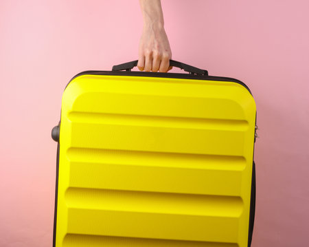 Hand holding Yellow plastic travel suitcase by handle on pink background. Tourism, vacation conceptの写真素材