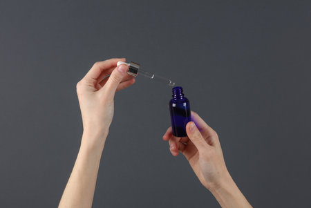 Hands holding serum bottle with pipette on dark gray background. Beauty concept