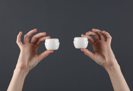 Hands hold white jars of cream on a dark gray background. Mockup, template for design, beauty...