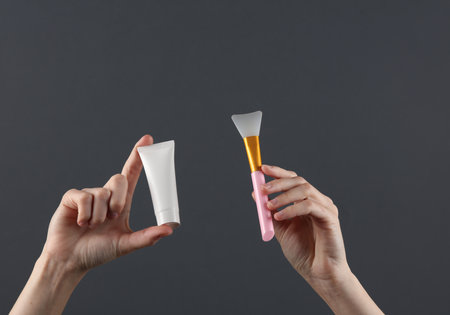 Woman's hand holds white cream tube and plastic brush on a dark gray background. Template for design, creative beauty mockupの写真素材