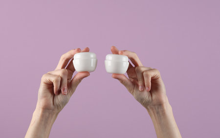 Hands holding white jars of cream on purple background. Mockup, template for design, beauty concept, skin careの写真素材