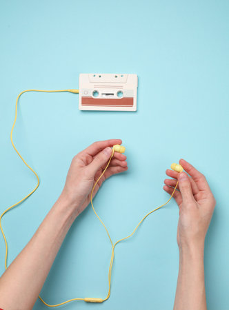 Hand holding wired headphones and audio cassette on blue background. Music lover, 80sの写真素材