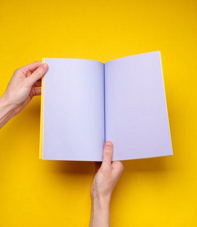 Female hands hold open notebook or magazine with purple blank pages on yellow backgroundの写真素材