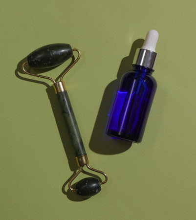 Massage jade roller and serum bottle on green background. Beauty concept