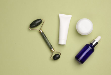 Massage jade roller and serum bottle, cream tube and jar on green background. Beauty concept. Flat...