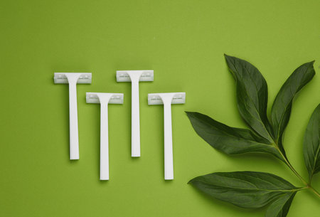 Disposable white plastic razors with green leaves on green background. Top view. Creative layout