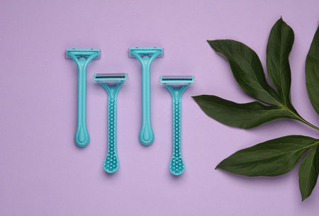 Disposable plastic razors with green leaves on purple background. Top view. Creative layout