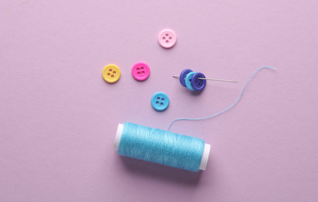 Skein of blue thread and a needle, buttons on lavender color background. Sewing accessories