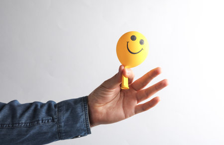 Man's hand in denim shirt holding Smiling face balloon on gray background.