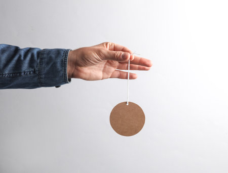 Man's hand in denim shirt holding craft round price tag on gray background. Sale conceptの写真素材