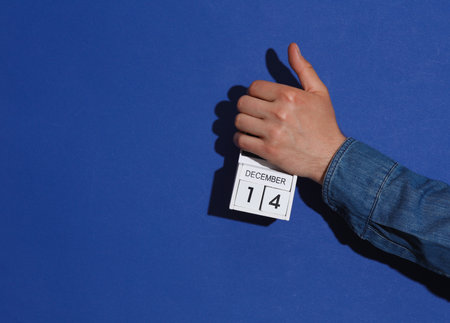 Man's hand in denim shirt holds wooden block calendar organizer with date December 14 on a blue backgroundの写真素材