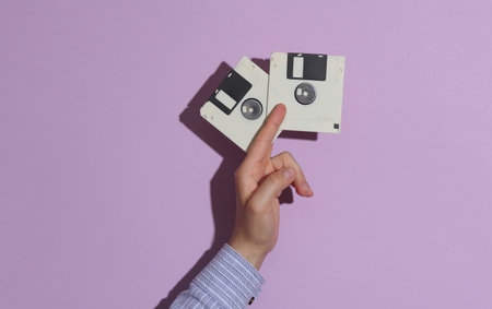 Man's hand in shirt holding two white floppy disks on purple pastel background with shadow