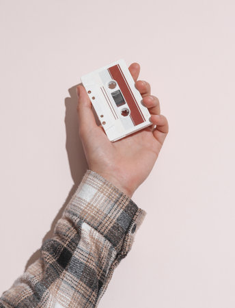 Man's hand in shirt holding audio cassette on beige background with shadow