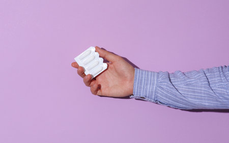 Man's hand in shirt holding rectal suppository for hemorrhoids on purple pastel background with...