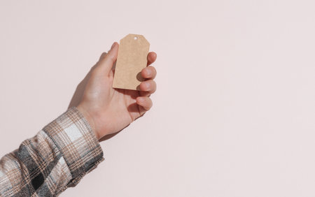 Man's hand in shirt holding blank craft price tag on beige pastel background with shadow. Sale...