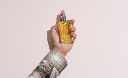 Man's hand in shirt holding perfume bottle on beige background with shadow
