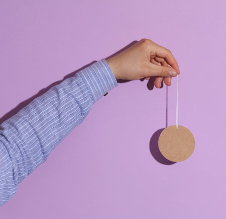 Man's hand in shirt holding blank craft round price tag on purple pastel background with shadow....