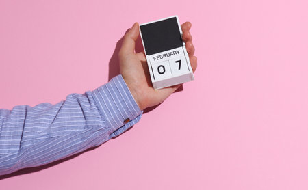 Man's hand in shirt wooden holds block calendar organizer with date February 7 on pink background...