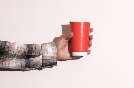 Man's hand in shirt holding red cardboard coffee cup on beige pastel background with shadowの写真素材
