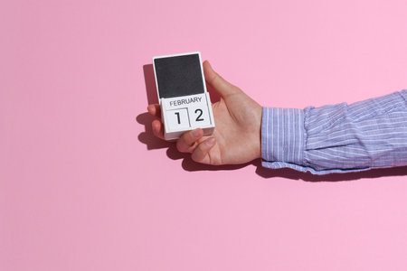 Man's hand in shirt wooden holds block calendar organizer with date February 12 on pink background with shadowの写真素材