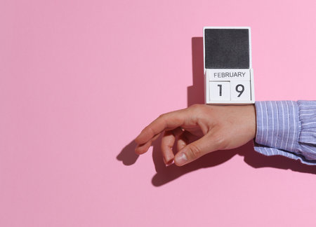 Man's hand in shirt wooden holds block calendar organizer with date February 19 on pink background with shadowの写真素材