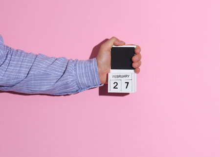 Man's hand in shirt wooden holds block calendar organizer with date February 27 on pink background with shadowの写真素材