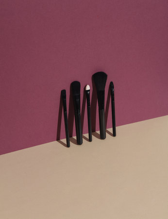 Black makeup brushes on burgundy beige background with shadow. Creative beauty layout.