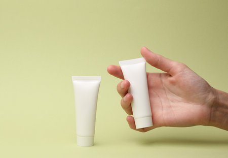 Hand holding White blank cream tube on green background. Template for design, mockup. Beauty conceptの写真素材