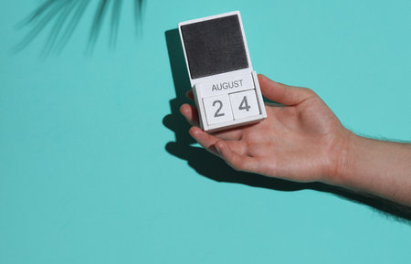 Man's hand holding wooden block calendar with date August 24 on blue background with palm leaf...