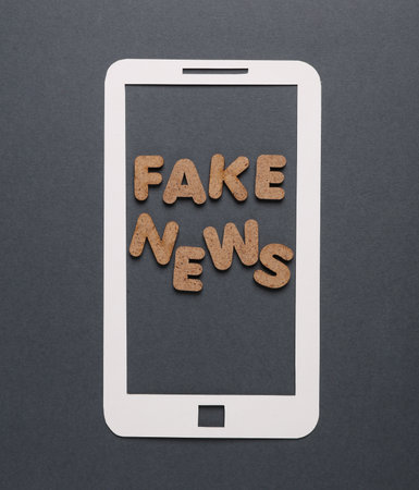 Paper cut smartphone with word fake news on dark gray background.